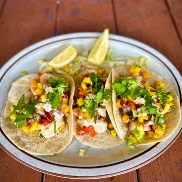 Three vibrant fish tacos with mango salsa, cilantro, and lime wedges on a plate.