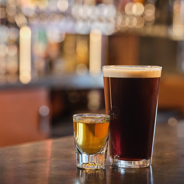 A dark beer with a foamy head next to a shot of amber liquor on a bar.