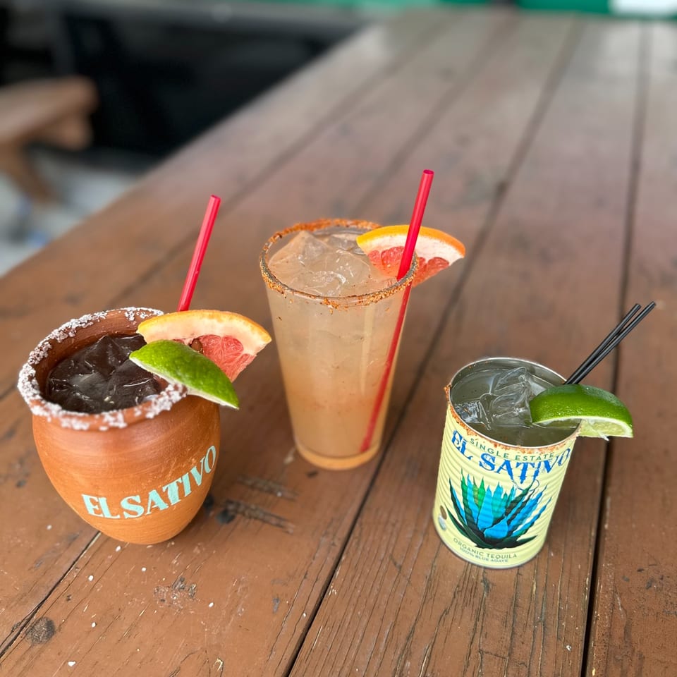 Three El Sativo tequila cocktails, two garnished with grapefruit and lime, on a rustic wooden table.
