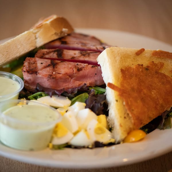 Grilled tuna steak salad with hard-boiled eggs, greens, and two dressing cups, served with toasted bread.