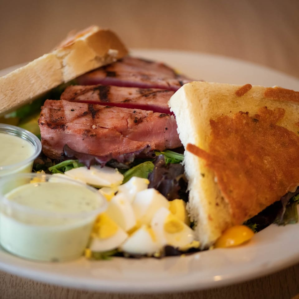 Grilled tuna steak salad with hard-boiled eggs, greens, and two dressing cups, served with toasted bread.