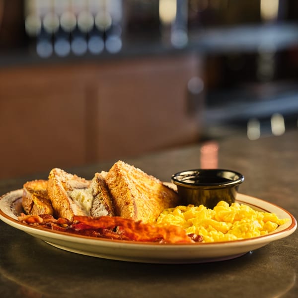 Legends Grill breakfast plate with French toast, scrambled eggs, bacon, and syrup on a dark counter.