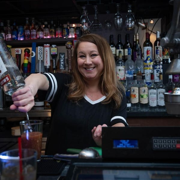 A smiling Legends Grill bartender pours a drink from a Smirnoff bottle behind a well-stocked bar.