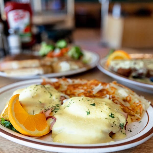 Eggs Benedict with hash browns and an orange slice, with other breakfast plates blurred in the background.