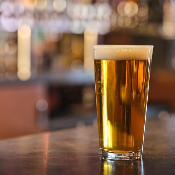 A pint glass of golden beer with a foamy head sits on a dark bar counter.